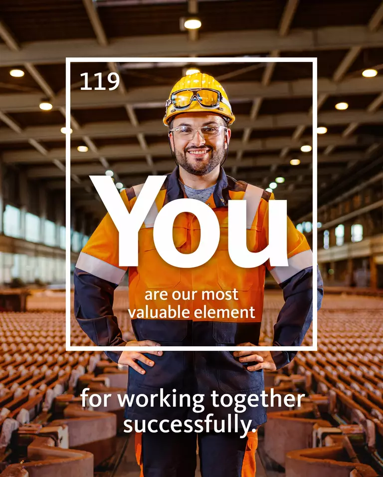 Smiling factory worker with hands on hips standing in a warehouse. Around him is a white frame with the words: ‘You are our most valuable asset in working together.’
