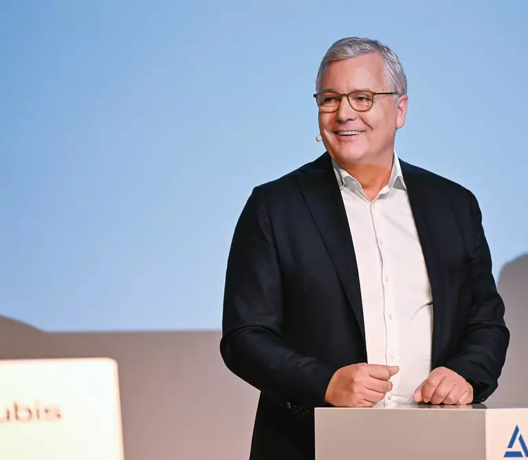 Dr. Toralf Haag, CEO, at a speaker´s podium wearing a black suit, white shirt, glasses and a name tag against a light blue background