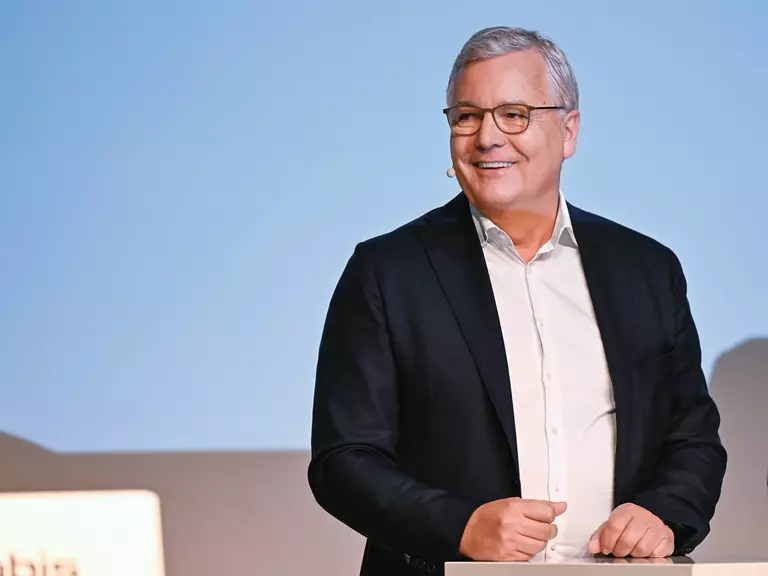 Dr. Toralf Haag at a speaker´s podium, wearing a black suit, white shirt, glasses and a name tag against a light blue background.