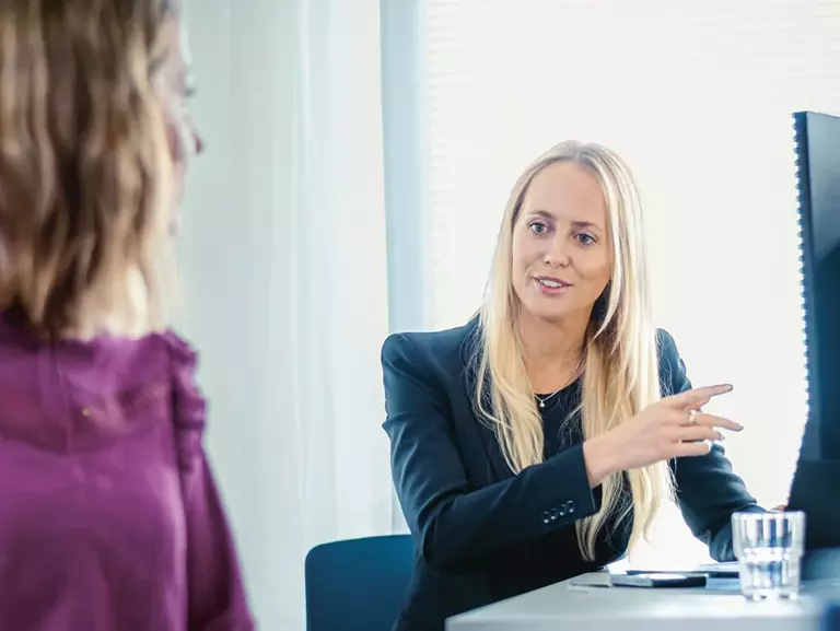 Conversation between two women, the first blonde in a black blazer, the second brunette in a pink top, at a table in an office.