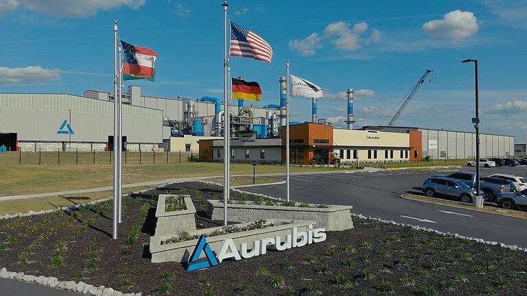 Video thumbnail showing Aurubis Richmond in the background. In the foreground, the Aurubis logo and several national flags, including those of Germany and the USA.