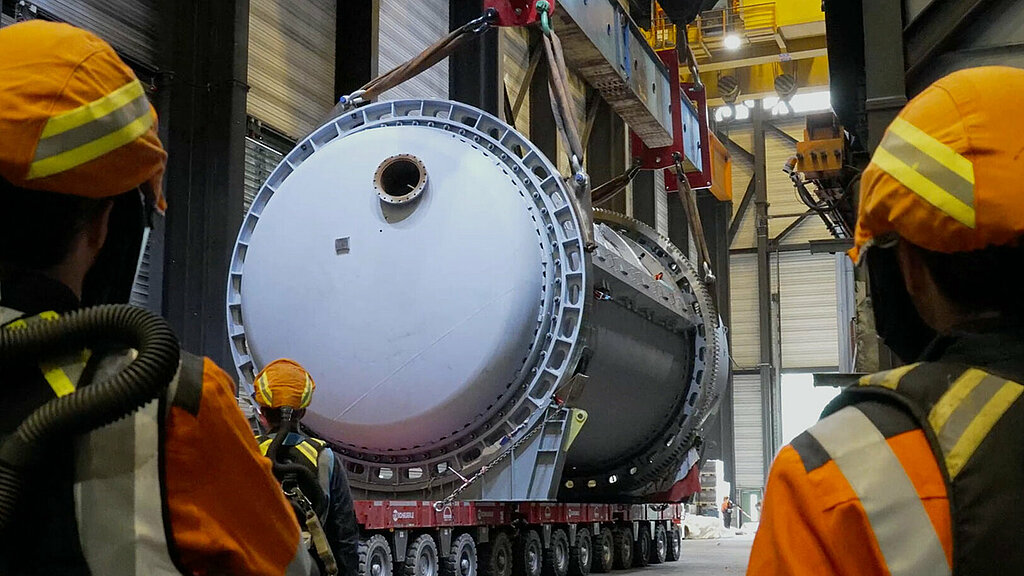 Two factory workers in protective clothing are seen from behind, standing in a factory hall in front of a crane device pulling a metal tube on rollers.
