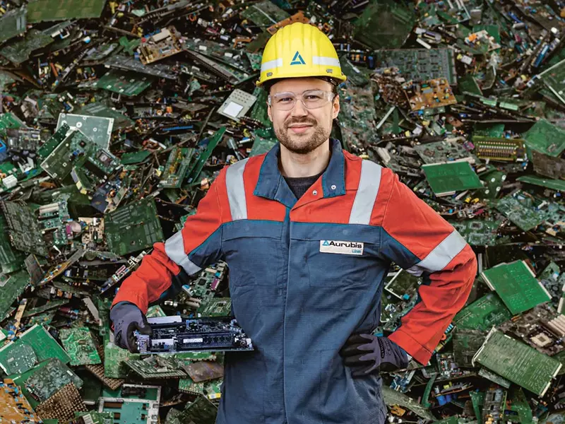 Tobias Kuhm, Executive Director of Supply Chain Management at Aurubis Lünen, standing in front of a pile of recyclable scrap metal wearing work clothes, safety goggles and a helmet