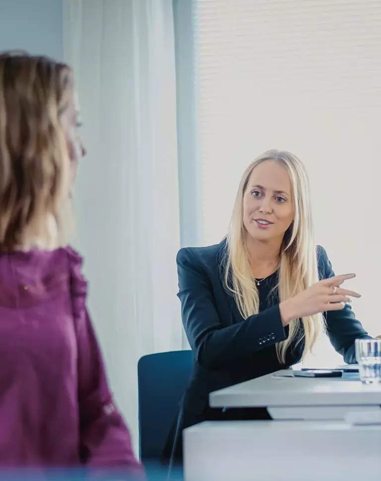 Conversation between two women, the first blonde in a black blazer, the second brunette in a pink top, at a table in an office