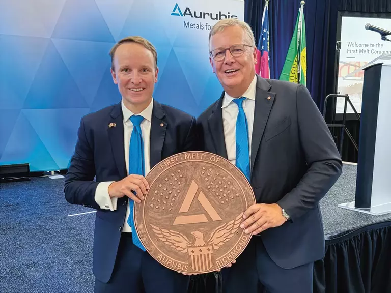 Two men in suits, white shirts and blue ties holding a round copper medal in their hands to mark the commissioning of the first US multi-metal recycling plant in Richmond. The medal features the Aurubis logo, with a stage in the background.