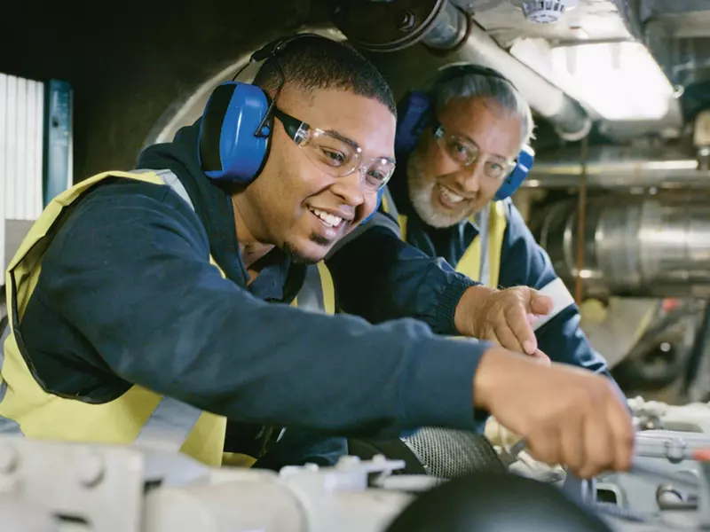 Two Aurubis employees wearing work clothes, safety goggles and ear protection working together. The older man is showing the younger man how to do a task.