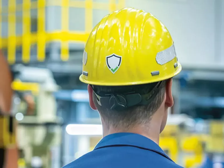 Back of the head of an employee wearing a yellow safety helmet; on the back, the new key visual of the Aurubis safety system.