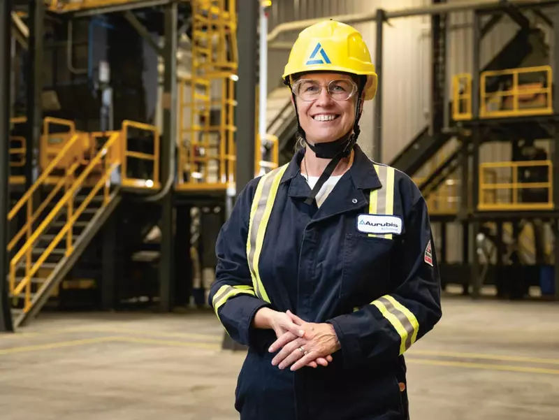 Inge Hofkens, COO, standing in a work hall wearing a yellow safety helmet and dark blue work clothes with reflectors on them