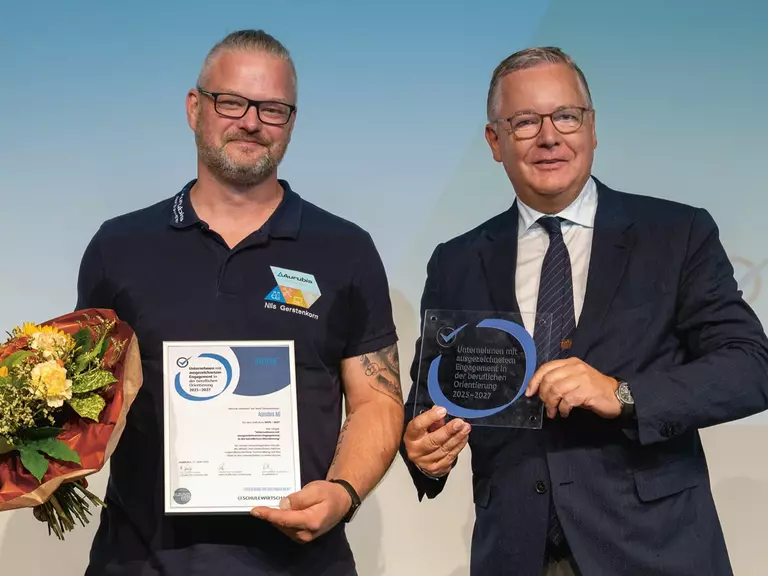 Two men on a stage holding a bouquet of flowers, a certificate and an award. Aurubis has been named one of Germany's best training providers for the fifth time, receiving the highest rating.