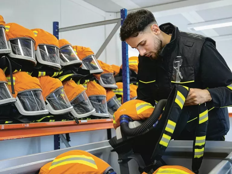 Young man in protective clothing lifting an orange respirator mask from a grey plastic box; behind him are two more rows of stacked masks.