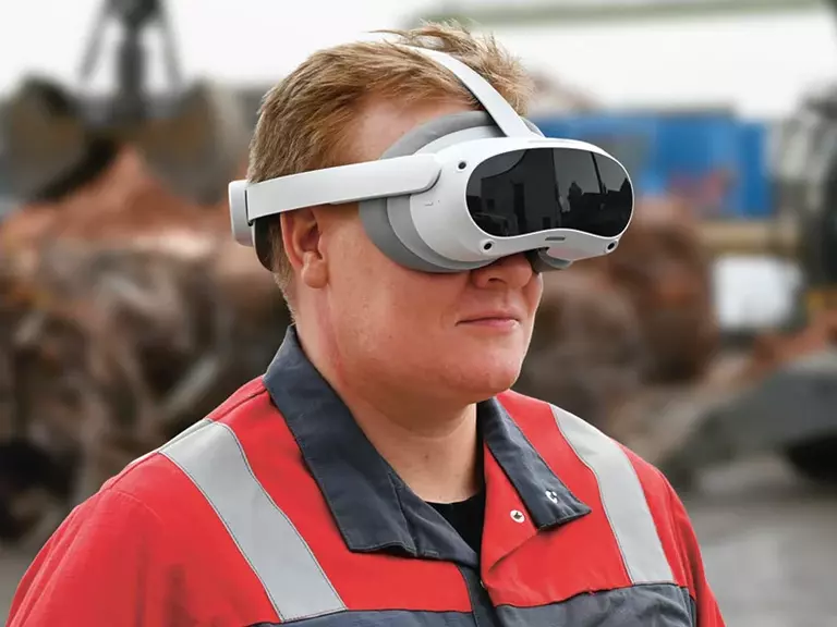 Man wearing work clothes and VR glasses on a construction site, with a crane bucket in the background.