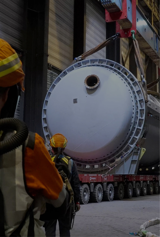 First level of Aurubis Performance 2030: Impact. Two factory workers in protective clothing are seen from behind, standing in a factory hall in front of a crane device pulling a metal tube on rollers.