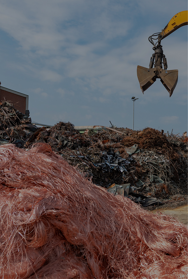Second level of Aurubis Performance 2030: Commercial Excellence. An excavator shovel is seen lowering onto a pile of scrap metal.